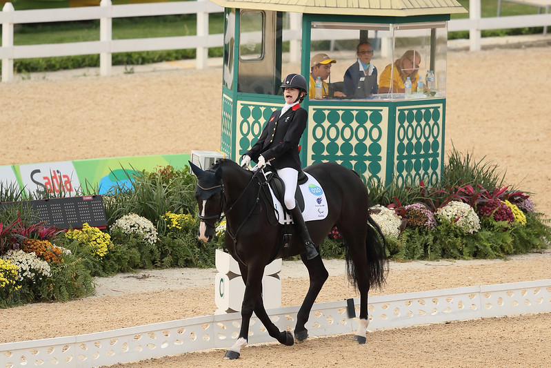 A woman riding a black horse. She is wearing a black riding jacket, black hat and white trousers. She is in front of a pavilion with people watching.