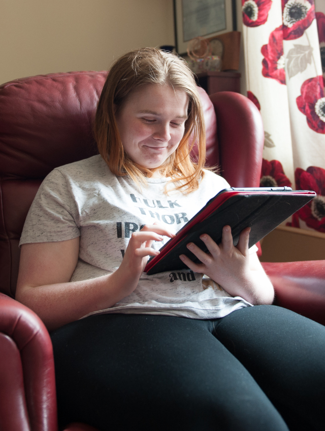 A smiling young woman sitting in a chair looking at a computer tablet device in her living room