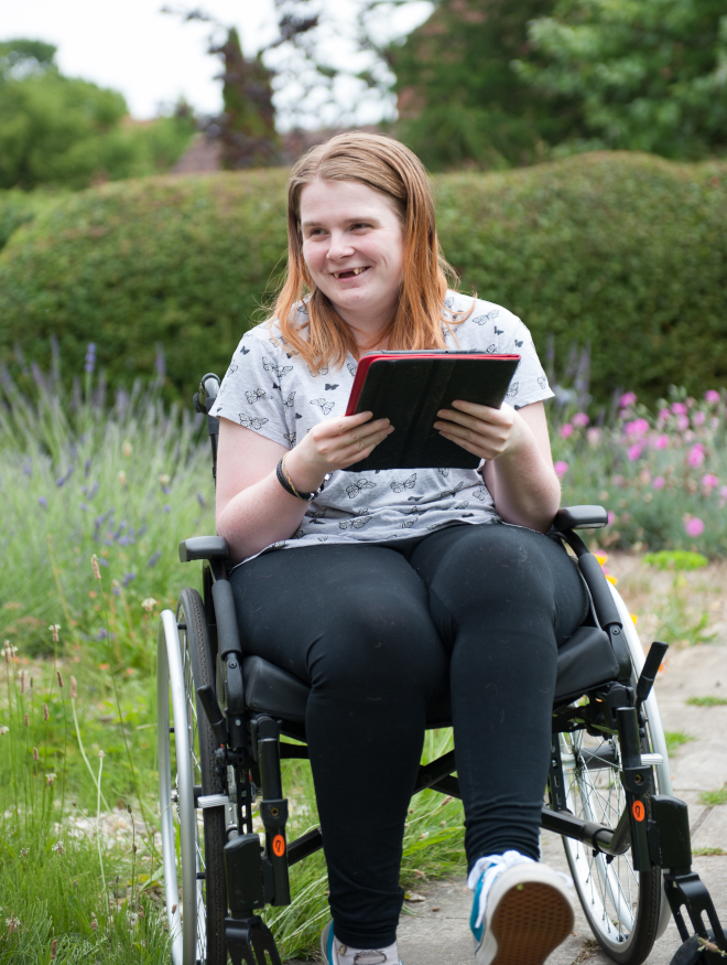 A smiling young woman outside in the garden in her wheelchair holding a computer tablet device