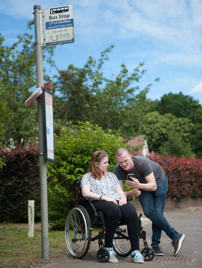 A man shows his phone screen to a woman in a wheelchair as they wait at a bus stop in a rural town