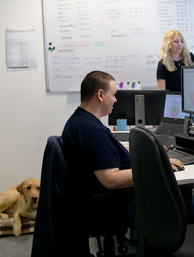 A man sat at a computer in an office. A dog sits on the floor next to him