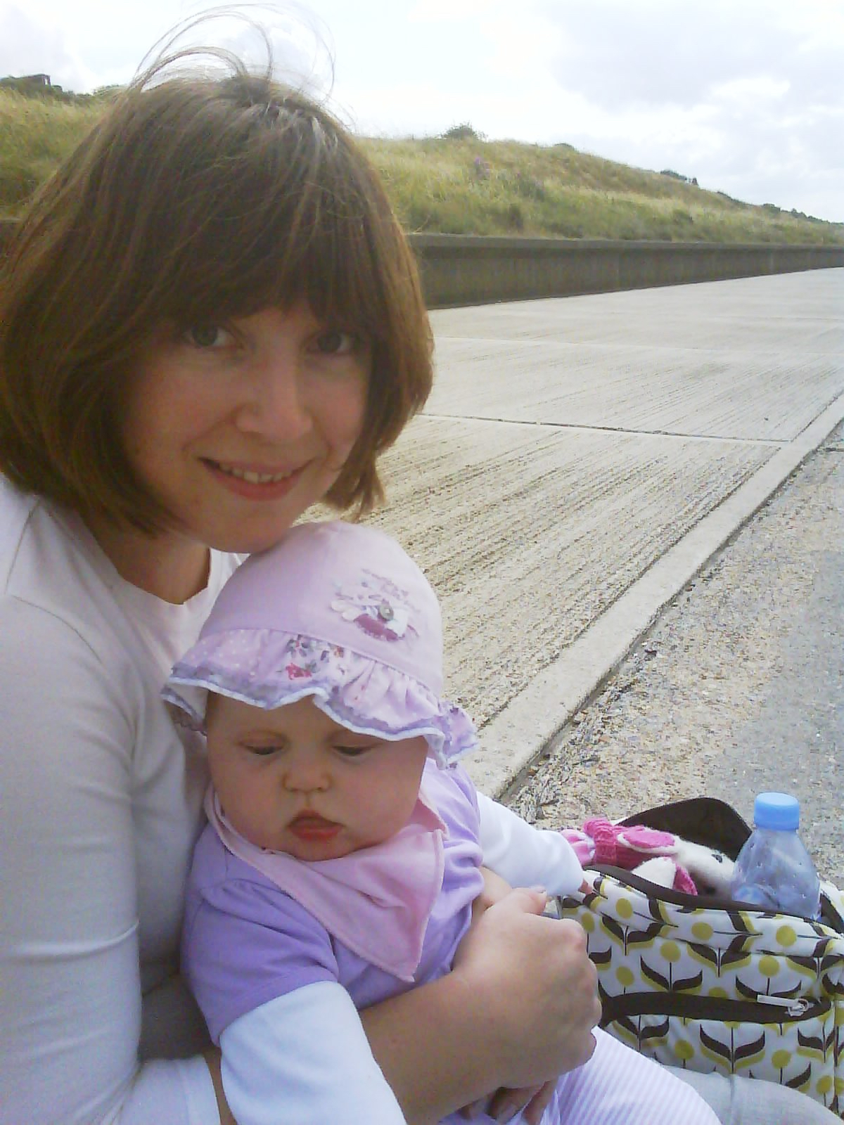 A woman and her baby daughter who is wearing a sun hat, sit outside near a grassy hill.