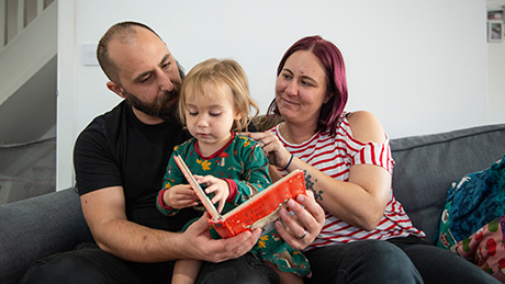Elara with her parents reading a book on the sofa looking happy.